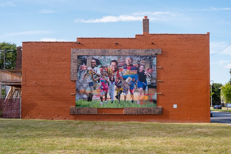 “Community Thrives Here” by Asia Hamilton features a group of children running playfully with a pair of elders, presumably grandparents, standing over them. 